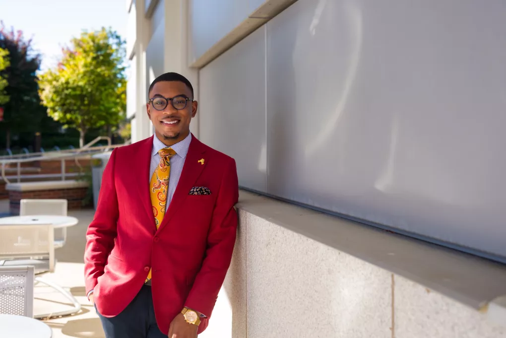 A young male sharply dressed in a red blazer and yellow tie poses for a photograph on UofL's campus.