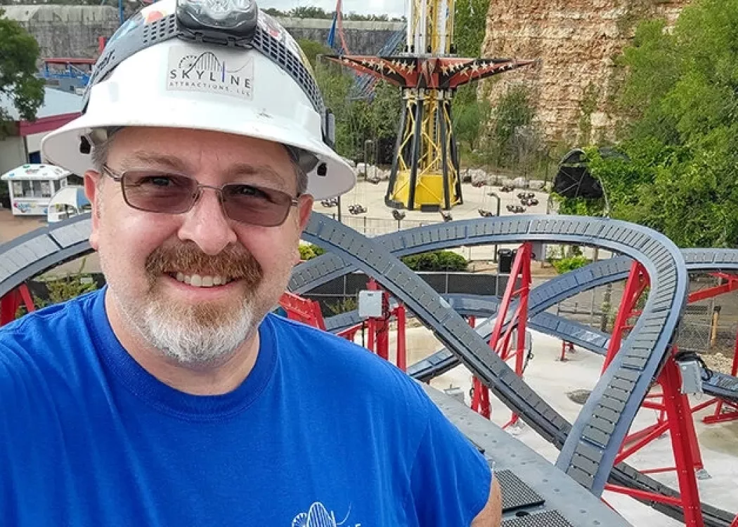 Alumnus Jeff Pike at an amusement park with a roller coaster behind him.