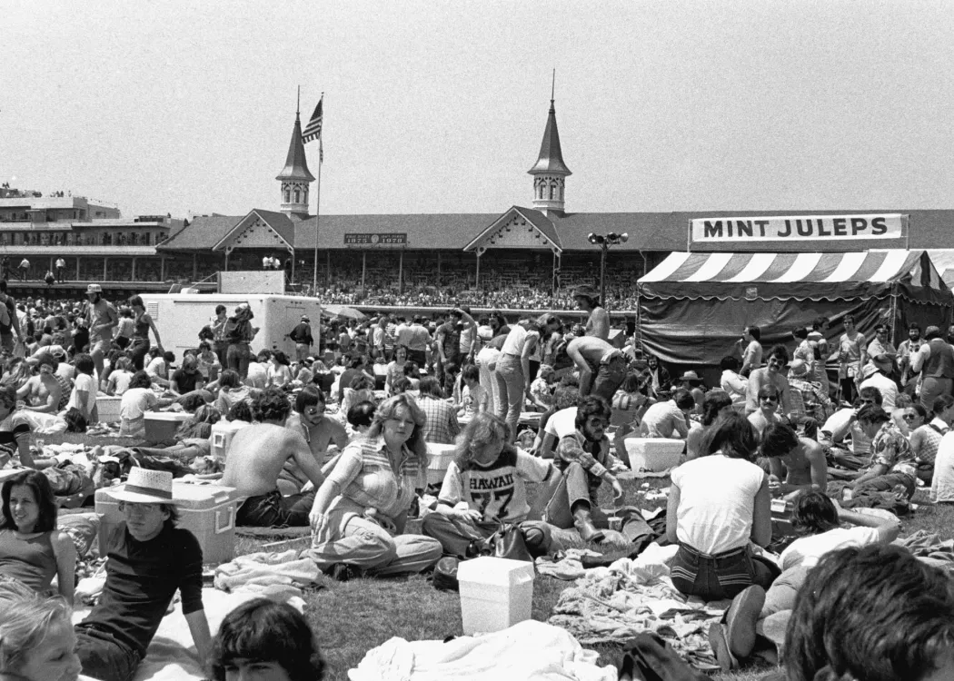 A black and white photo of the infield crowd at the 1978 Kentucky Derby. The crowd is gathered around a tent selling mint juleps, with the historic Twin Spires behind it.