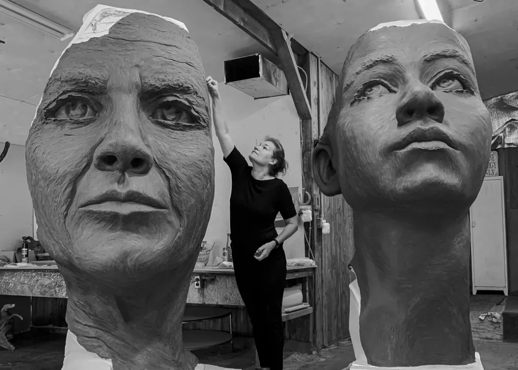 a woman is carving and surrounded by giant faces created out of clay in an artist studio.