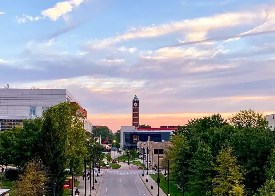 A sunrise over UofL creating a pink, yellow, and blue morning sky. UofL's clock tower and student union can be seen in the background amongst green trees.