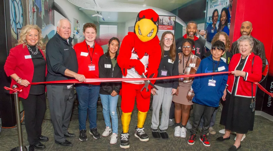 a group of men, women and children join a mascot dressed as a Cardinal to cut a ribbon