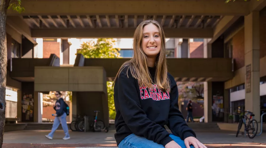 A young woman wearing a black sweatshirt with Louisville Cardinals in red lettering on the front poses for a picture on the UofL campus quad.