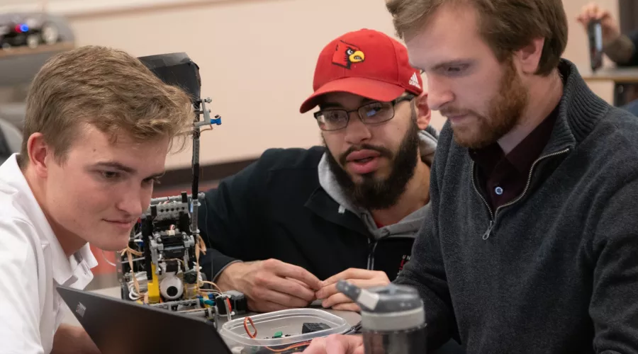 Three electrical and computer engineering students looking at a laptop alongside the small robot they made to compete in their class final.